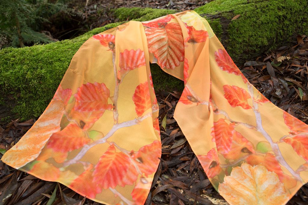Van Diemen Designs Fagus scarf draped over a mossy log, in orange and red autumn tones